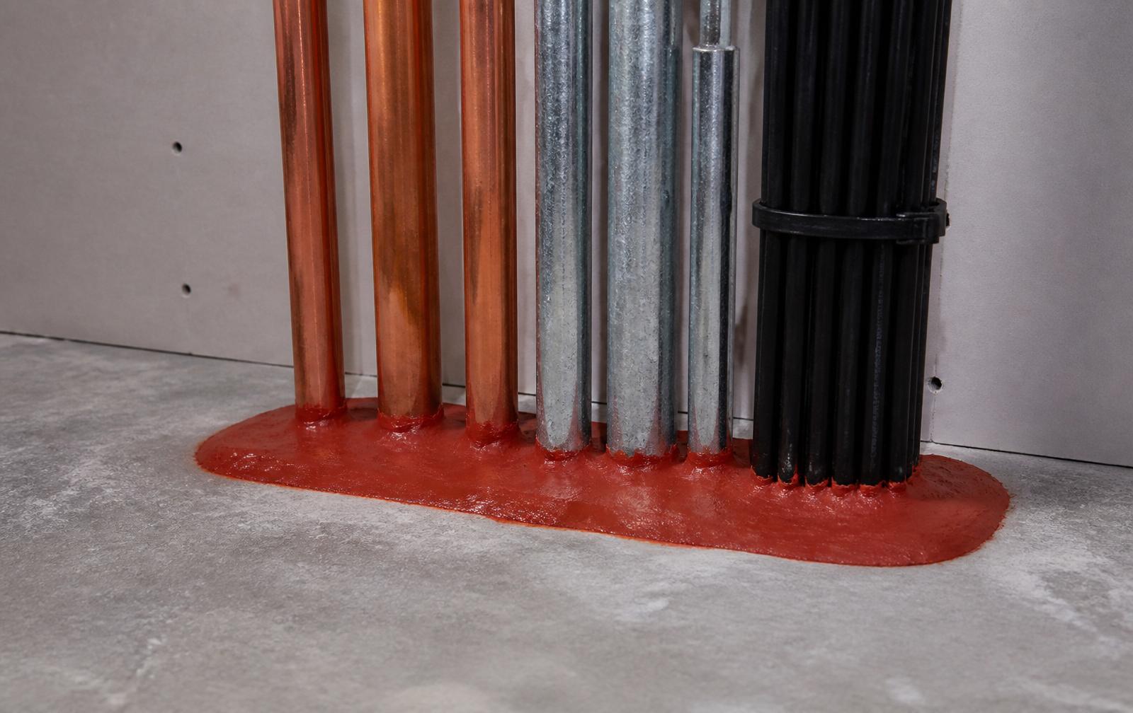 Red intumescent fire stopping compound sealed around copper pipes and cables penetrating a floor slab