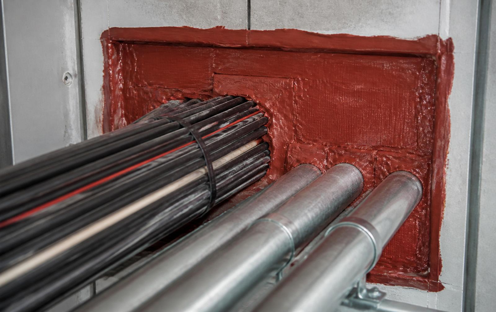Service penetration sealed with red intumescent mastic around cables and conduits through a compartment wall